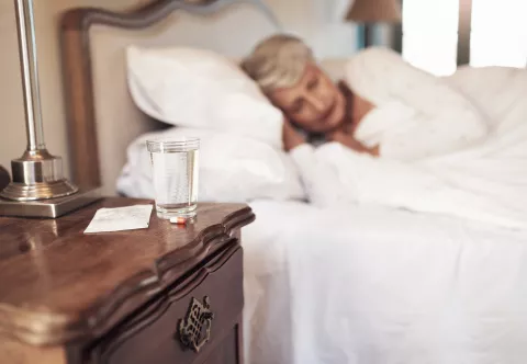 Medicijnen en een glas water met op de achtergrond een oudere vrouw slapend in haar bed